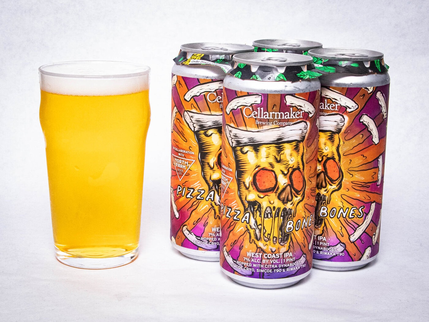 A Pint Glass Of Hazy Golden Beer Sits Next To A Four-Pack Of Colorful Cellarmaker &Quot;Pizza Bones&Quot; West Coast Ipa Cans, Featuring A Graphic Of A Skull With Pizza Slices And Bones On The Label.