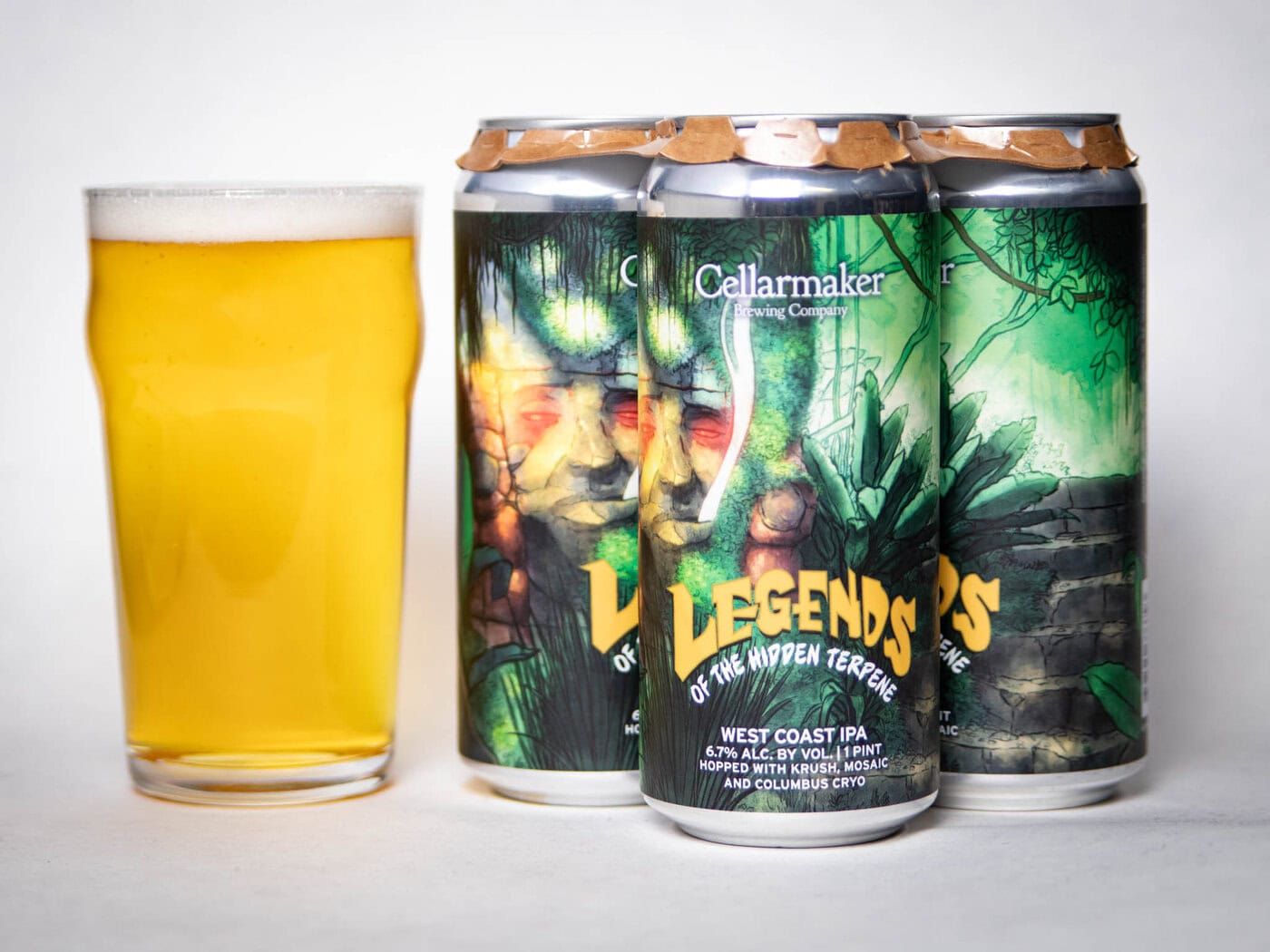 A Glass Of Hazy Yellow Beer Stands Next To Three Cans Of Cellarmaker &Quot;Legends Of The Hidden Terpene&Quot; West Coast Ipa, Featuring Vibrant Green, Jungle-Themed Artwork On The Cans.