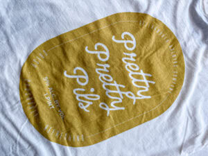 The Pretty Pretty Pils Tee is a white shirt with a large yellow oval label that reads "Pretty Pretty Pils" in white script, plus “5% ALC. BY VOL. 1 PINT,” “Crisp,” “Refreshing,” and “Commonwealth Brewing Company.”.
