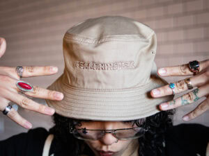 A person wearing the Cellarmaker Bucket Hat, embroidered with "DREAMERMAKER," sports rings on both hands and glasses, standing before a background of light-colored tiles.