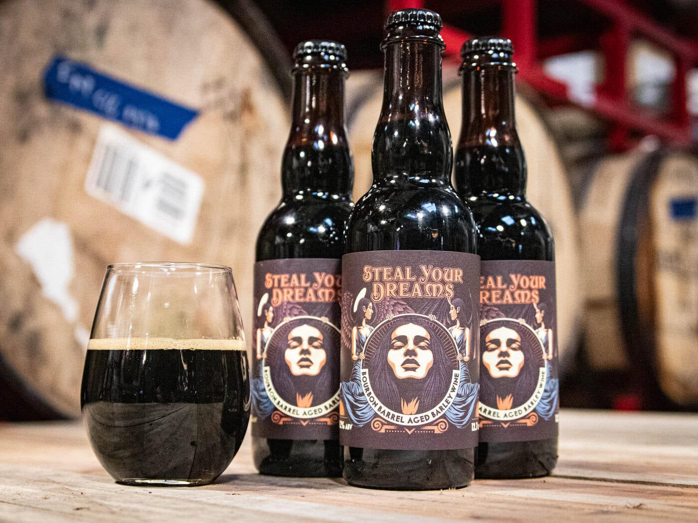 Three Bottles Of &Amp;Quot;Steel Your Dreams - Ba Barleywine&Amp;Quot; With Illustrated Labels Stand On A Wooden Surface Beside A Glass Of Dark Beer; Large Barrels Appear Blurred In The Background.