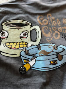 The Pretty Pretty Pils Tee features a cartoon coffee cup with yellow teeth and wide eyes beside an annoyed blue ashtray with a cigarette. "Coffee & Cigarettes" is partially visible in orange lettering on the fabric.