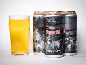 A pint glass filled with pale yellow beer sits next to three cans labeled "Cellarmaker Super Hi! Japanese Style Lager" with a dark, cloudy design on the cans.