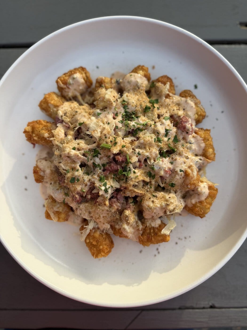 A White Plate With Crispy Tater Tots Topped With A Creamy Sauce, Ground Meat, Shredded Cheese, And Chopped Herbs, Served On A Gray Wooden Table.