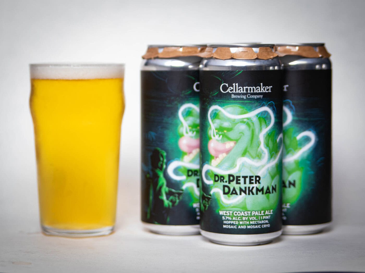 A Pint Glass Of Hazy Yellow Beer Sits Beside Four Cans Of &Quot;Christopher Riwakan - Hazy Pale Ale&Quot; From Cellarmaker Brewing Company, Each Can Decorated With A Green Cartoon Ghost.