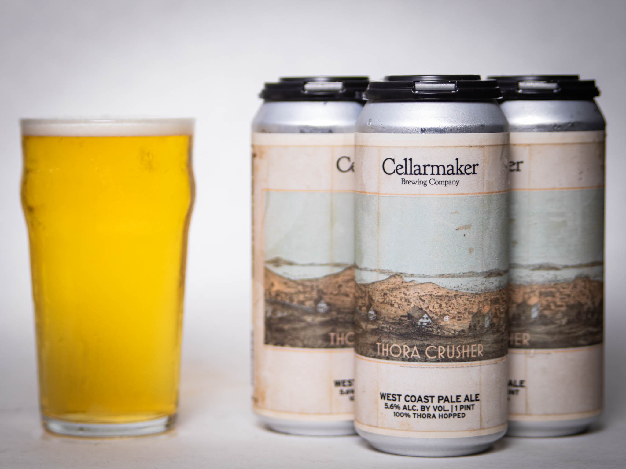 A Pint Glass Of Hazy Pale Yellow Beer Sits Beside Three Cans Of Cellarmaker Brewing Company’s Christopher Riwakan Hazy Pale Ale, All Set Against A Plain White Background.