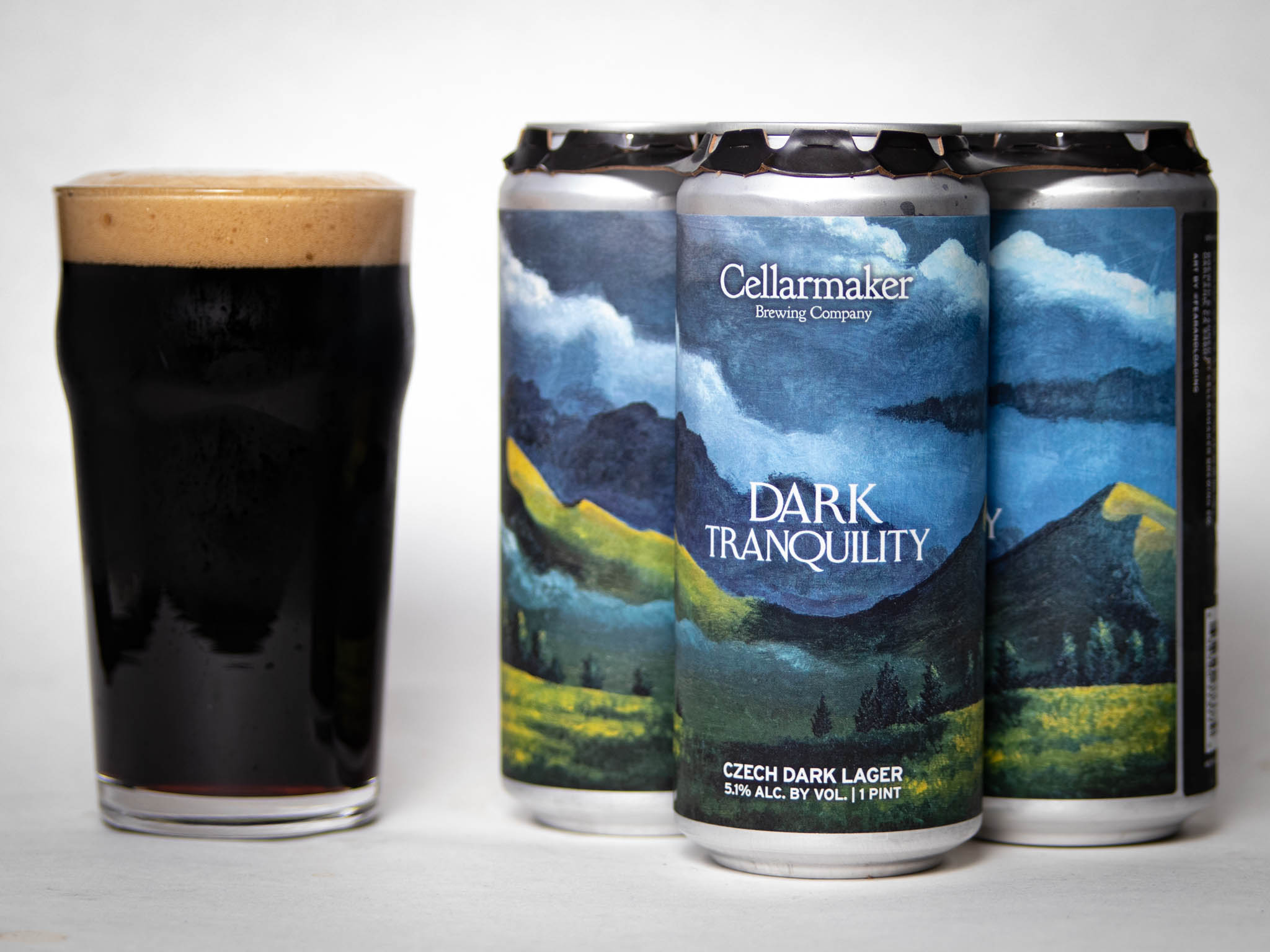 A Pint Glass Filled With Dark Beer Sits Next To Three Cans Of Cellarmaker Brewing Company’s Dark Tranquility Czech Dark Lager, Featuring A Mountain Landscape Design On The Labels.