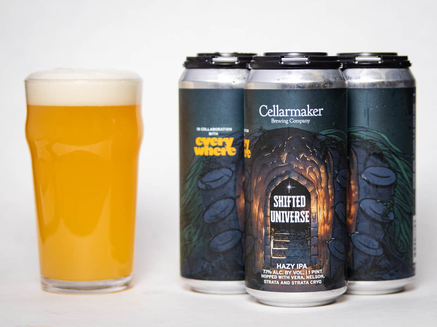 A Pint Glass Of Hazy, Golden Beer Sits Next To Four Cans Labeled &Quot;Shifted Universe Hazy Ipa&Quot; By Cellarmaker Brewing Company. The Cans Feature Dark, Illustrated Tunnel Artwork With Glowing Light At The Center.
