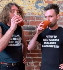 Two men stand by a brick wall, both wearing Merel Haggard Shirts and drinking reddish beverages with the Cellarmaker Beer logo visible on their shirts.