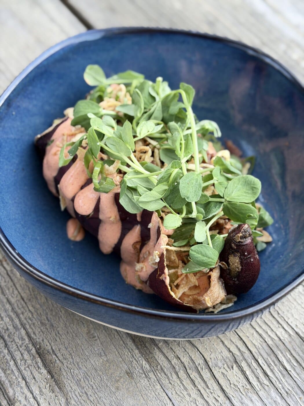 A Baked Sweet Potato Topped With A Pink Sauce, Crispy Fried Onions, And Fresh Green Sprouts, Served In A Blue Bowl On A Rustic Wooden Table.