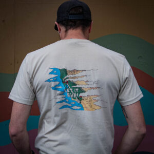 A person wearing the Let it Riip Shirt—a beige tee with a colorful graphic and "LET IT RIP!" text on the back—stands facing a painted mural. They also wear a black cap and earrings.