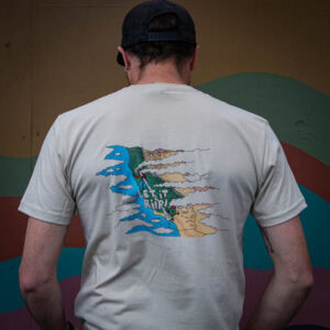 A person wearing the Let it Riip Shirt—a beige tee with a colorful graphic and "LET IT RIP!" text on the back—stands facing a painted mural. They also wear a black cap and earrings.