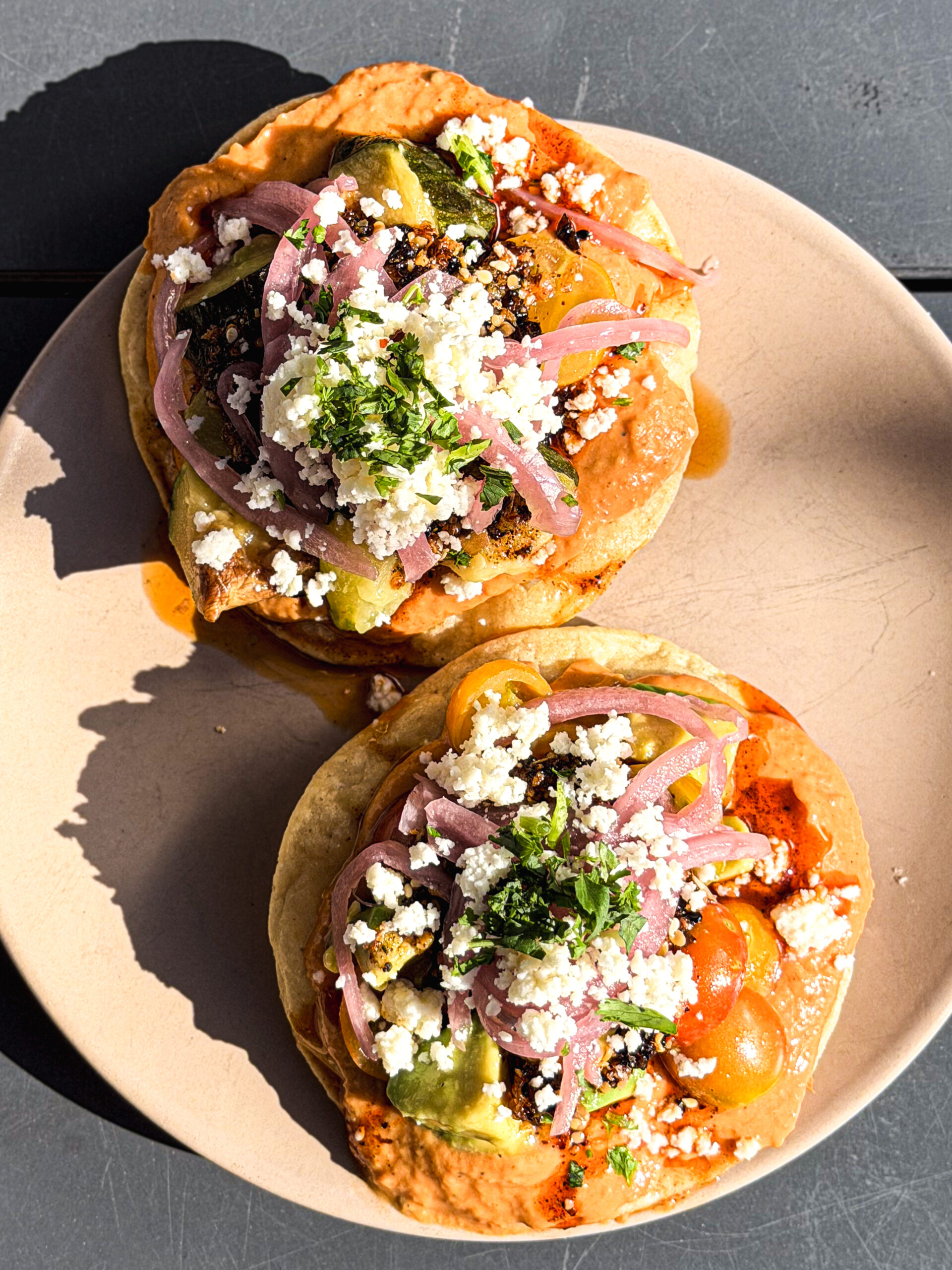 Two Crispy Tostadas Topped With Colorful Ingredients Including Crumbled Cheese, Pickled Onions, Chopped Herbs, And A Mix Of Vegetables, Served On A Light Tan Plate. Sunlight Casts Bright Highlights Across The Dish.