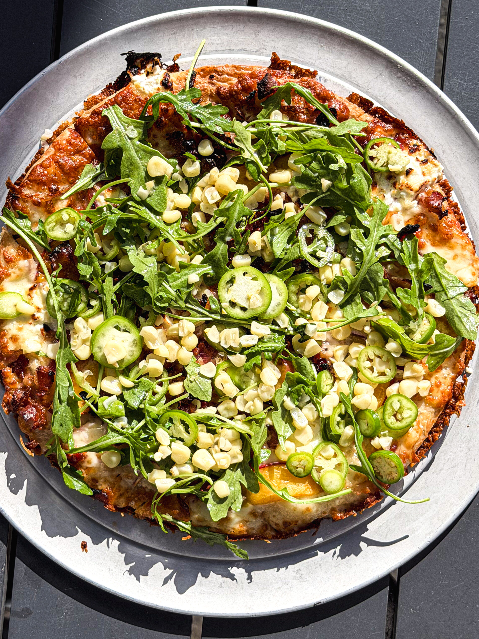 A Pizza Topped With Fresh Arugula, Sliced Green Peppers, Corn Kernels, And Melted Cheese Sits On A Round White Plate On A Dark Table.