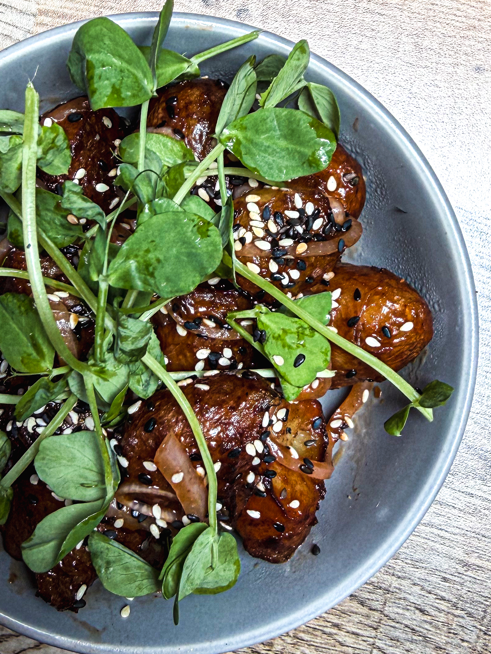 A gray bowl filled with roasted baby potatoes topped with fresh greens, black and white sesame seeds, and thinly sliced onions, sitting on a light wooden table.