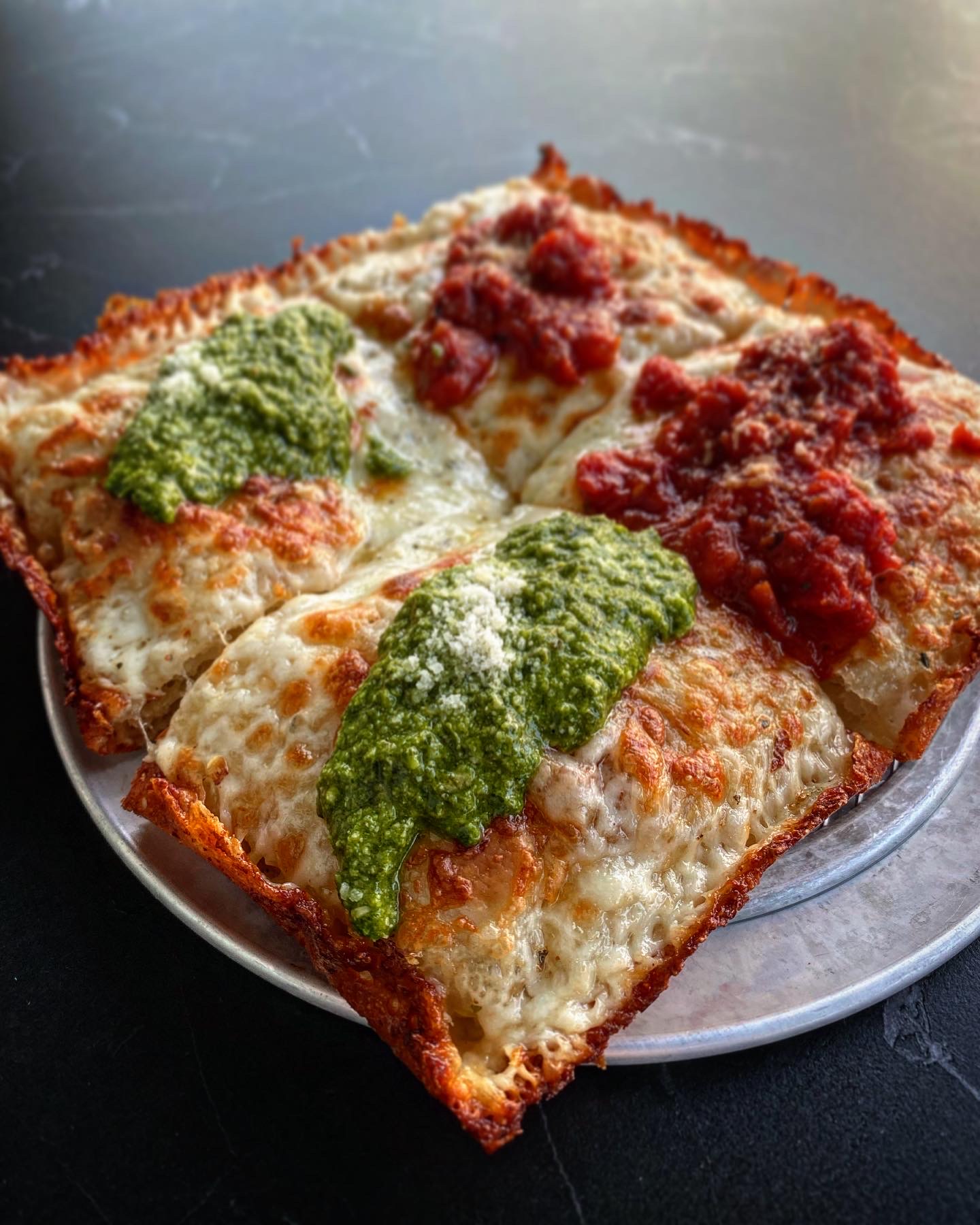 A close-up of four square slices of Detroit-style pizza on a round metal tray, topped with melted cheese, tomato sauce, and green pesto sauce, set against a dark background.