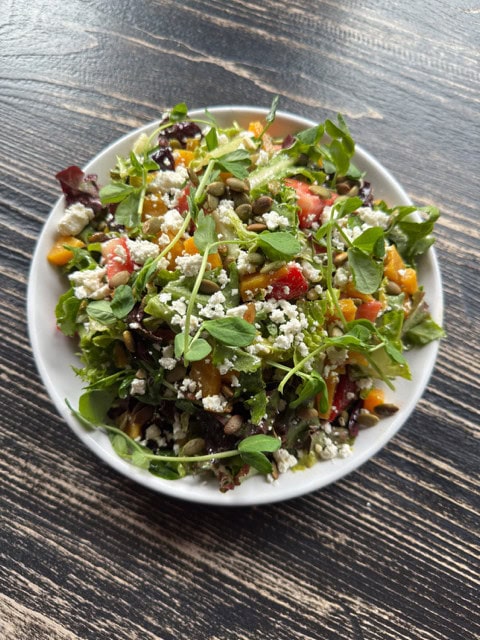 A white bowl filled with a colorful salad topped with crumbled cheese, seeds, and microgreens, sitting on a rustic wooden table.