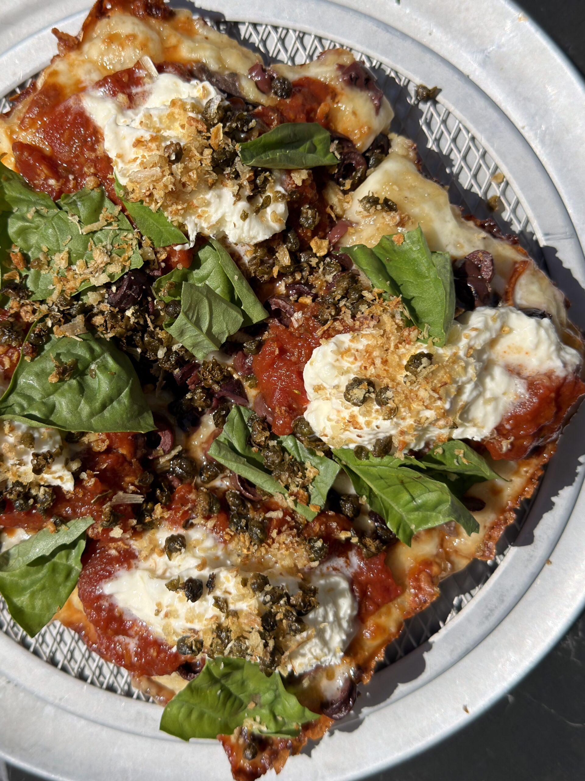 A close-up of a plate with slices of pizza topped with tomato sauce, melted cheese, dollops of ricotta, fresh basil leaves, and a crispy breadcrumb garnish. The pizza is served on a wire rack atop a round white plate.