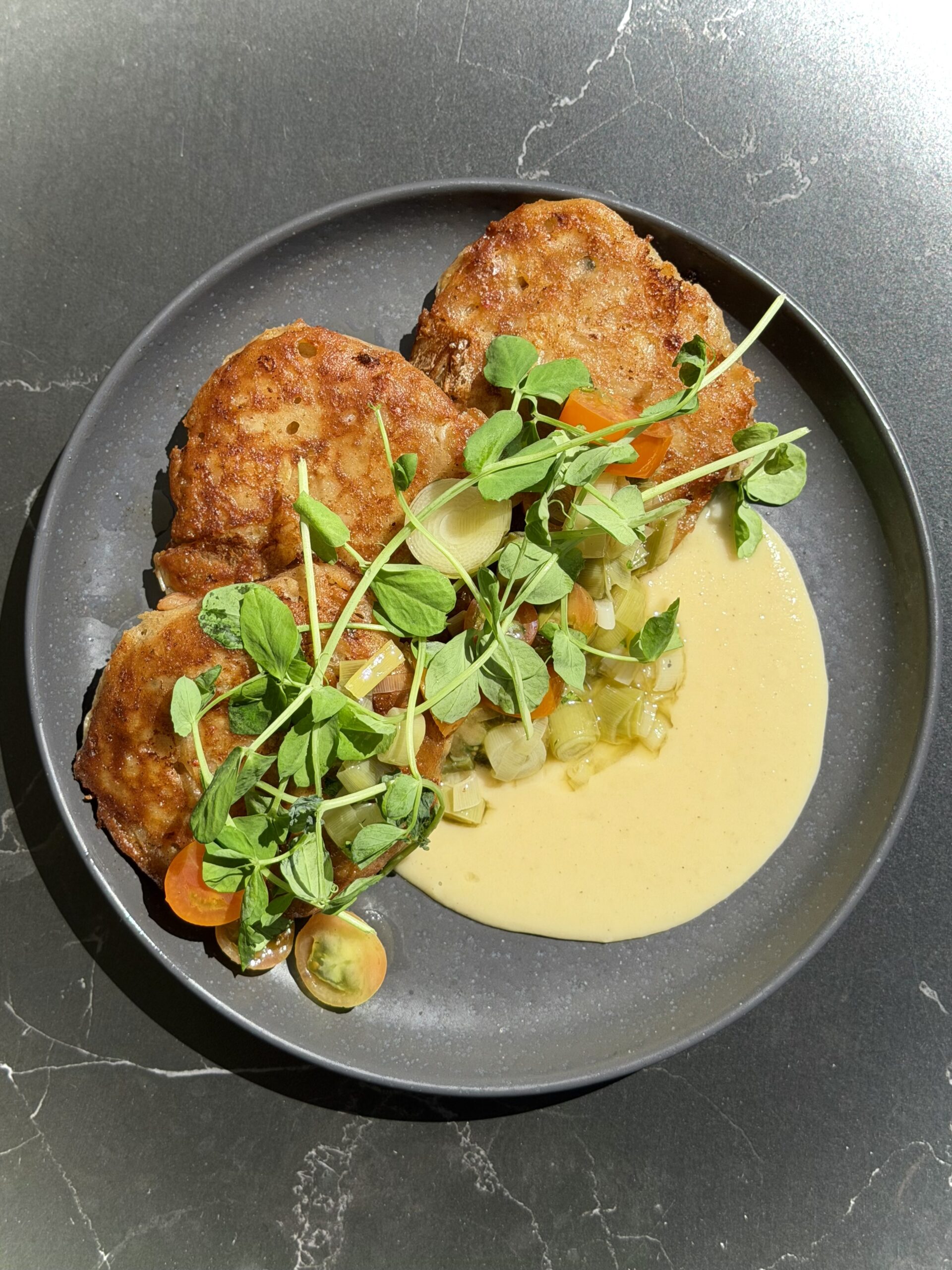 A dark plate with three golden-brown fritters, fresh microgreens, sliced vegetables, diced pickles, and a creamy yellow sauce, served on a dark marble surface.