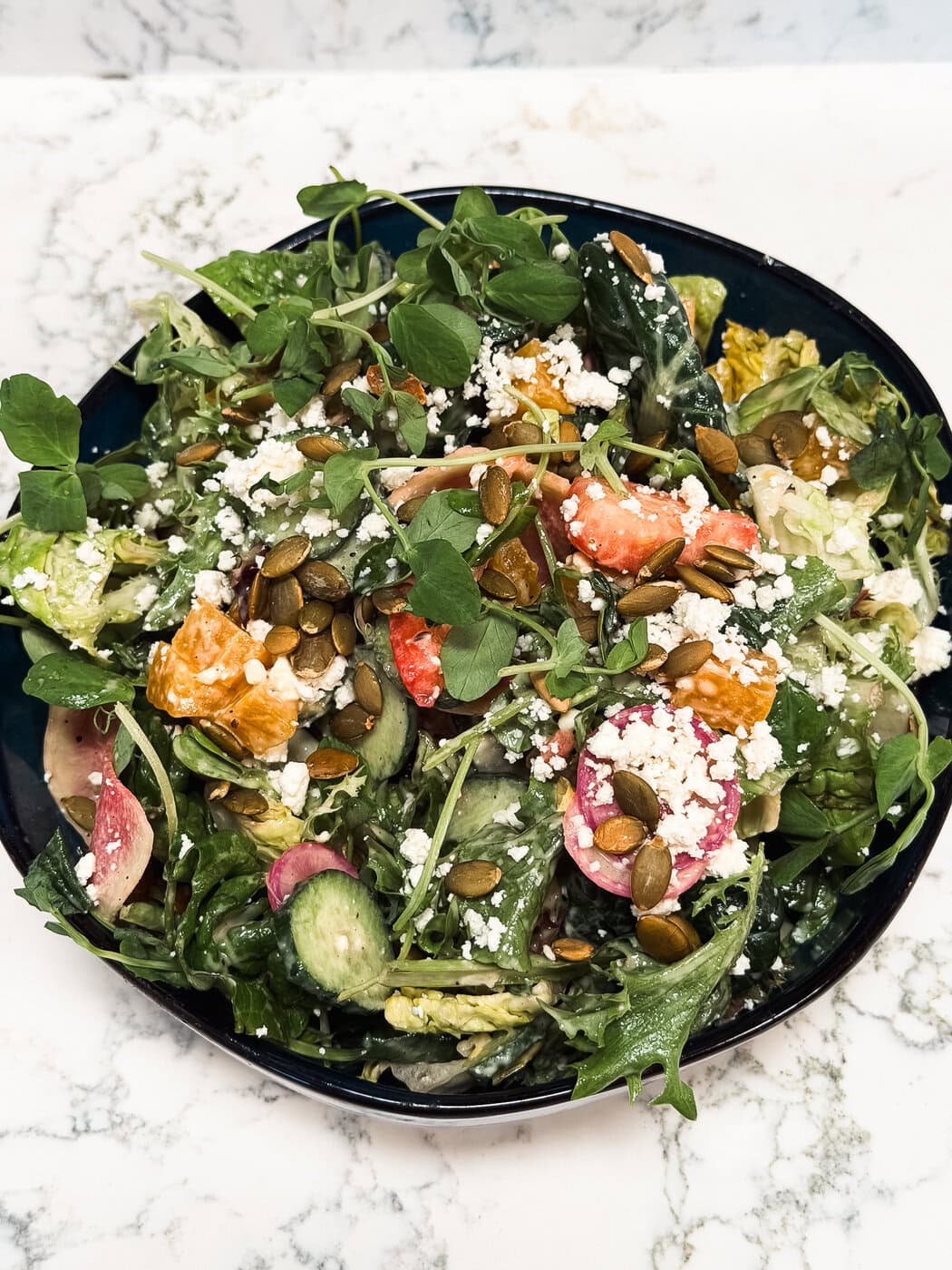 A bowl of mixed green salad with feta cheese, sliced radishes, cucumbers, tomatoes, orange segments, and pumpkin seeds on a marble surface.
