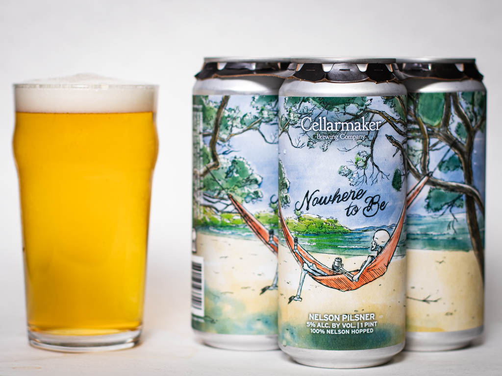 A Pint Glass Filled With Pale Beer Sits Beside Four Cans Labeled “Cellarmaker Brewing Company Nowhere To Be,” Featuring Artwork Of A Person Relaxing In A Hammock On A Beach Under Trees.