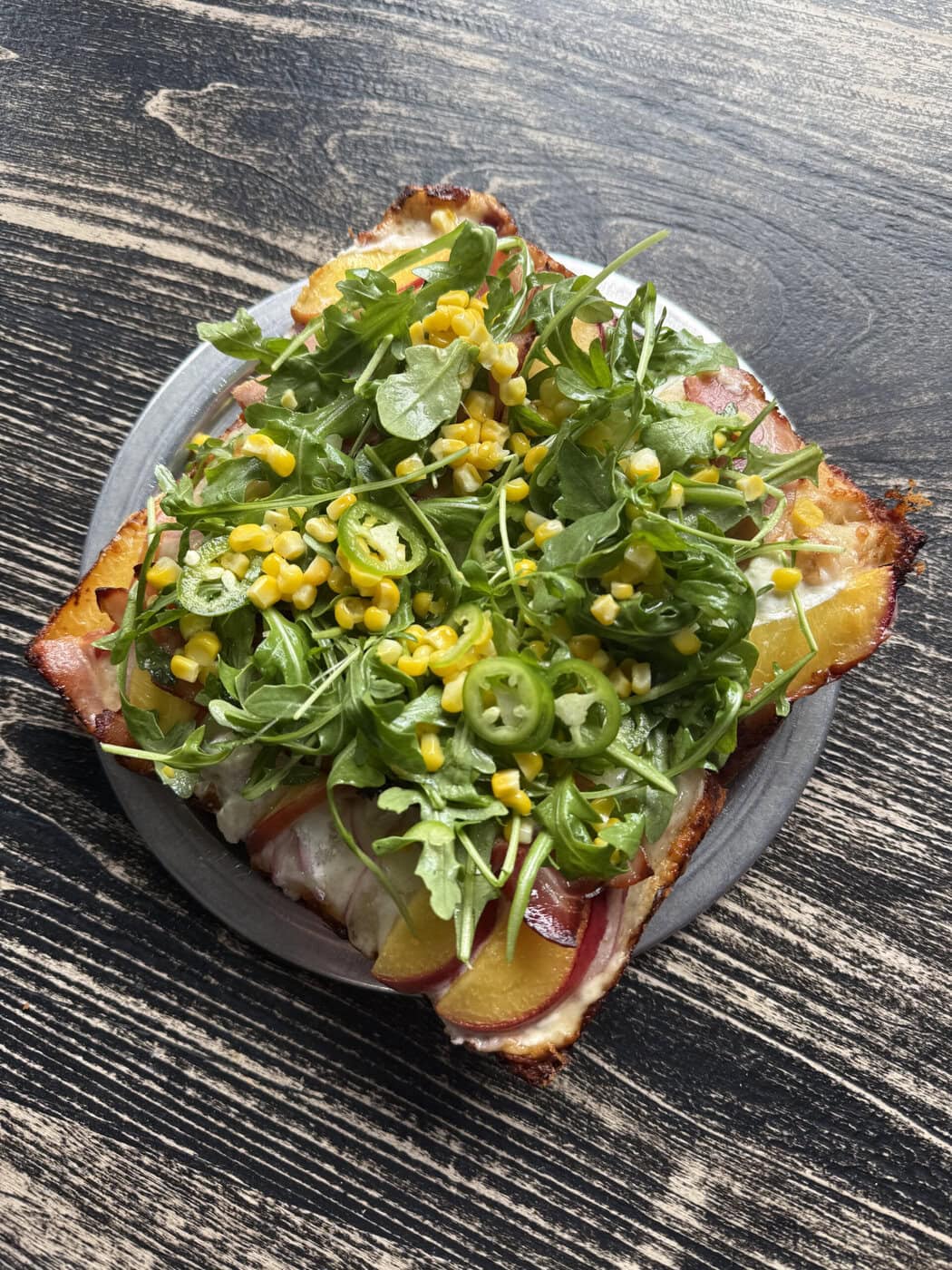 A Square Slice Of Pizza Topped With Melted Cheese, Sliced Peaches, Arugula, Corn, Sliced Green Peppers, And Red Onions Sits On A Gray Plate Atop A Textured Dark Wood Table.