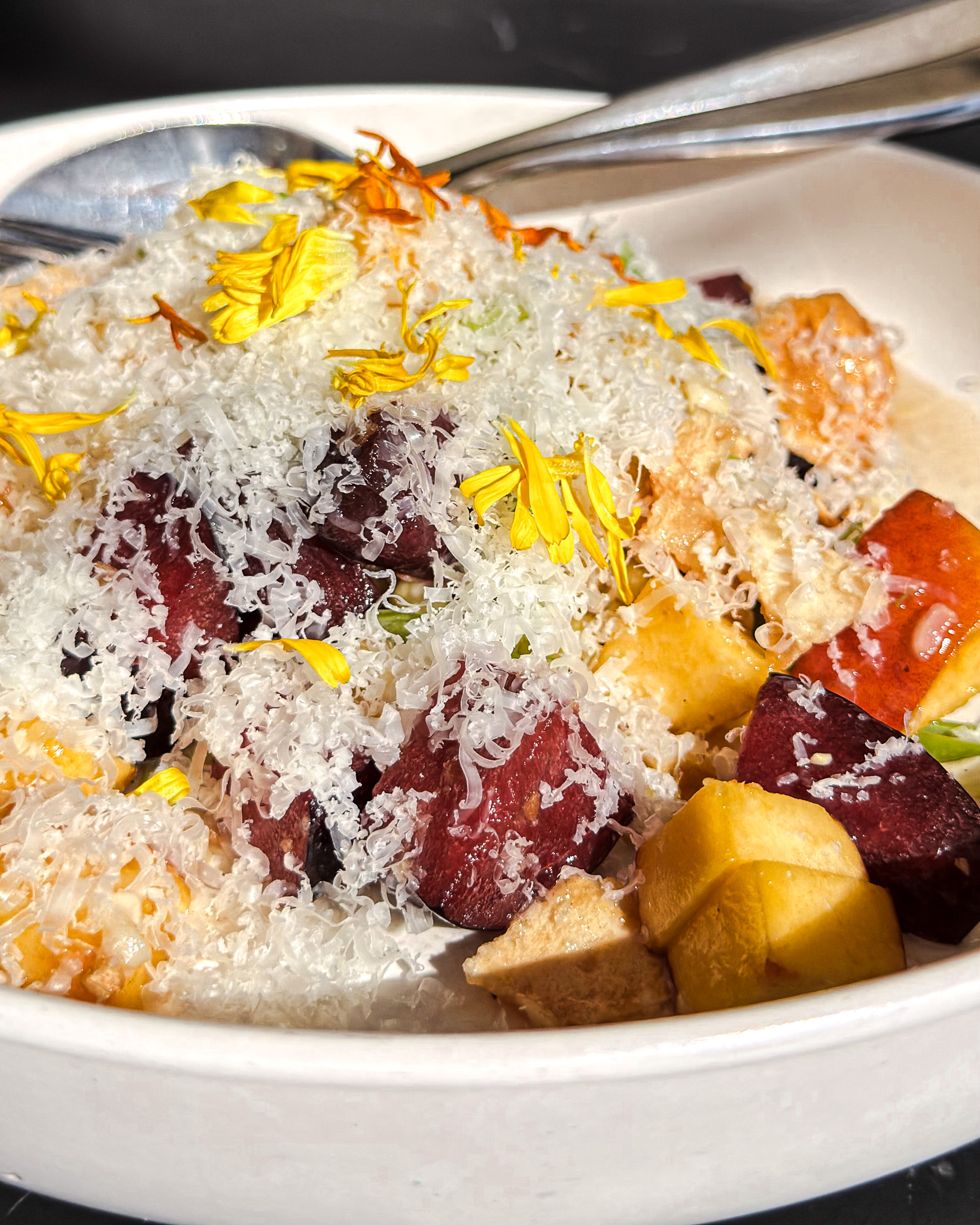 A Close-Up Of A Salad In A White Bowl With Diced Beets, Yellow Flowers, Cheese Shavings, And Mixed Vegetables, With Two Metal Spoons In The Background.
