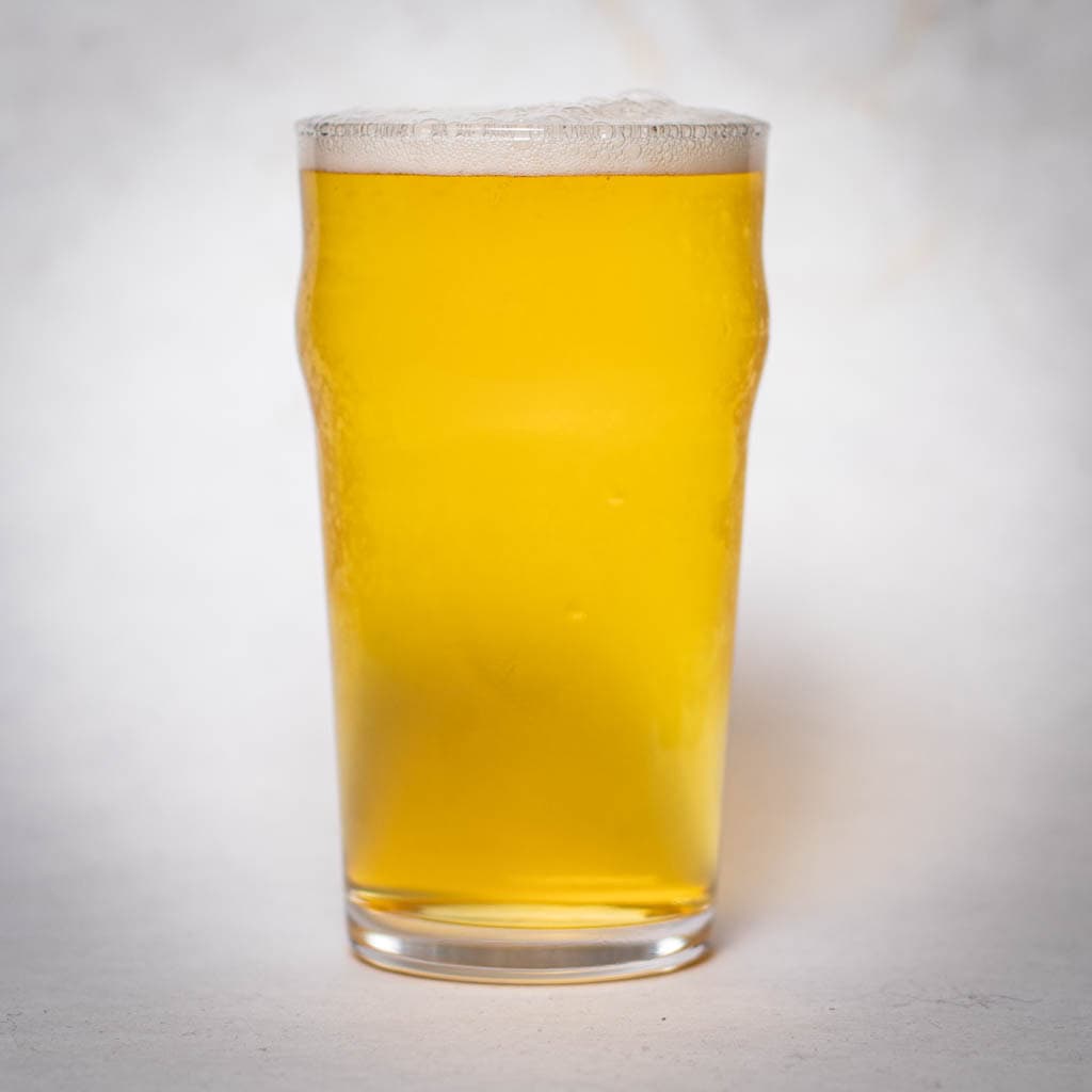 A Clear Pint Glass Filled With Golden-Yellow Beer, Topped With A Thin Layer Of Foam, Set Against A Plain White Background.