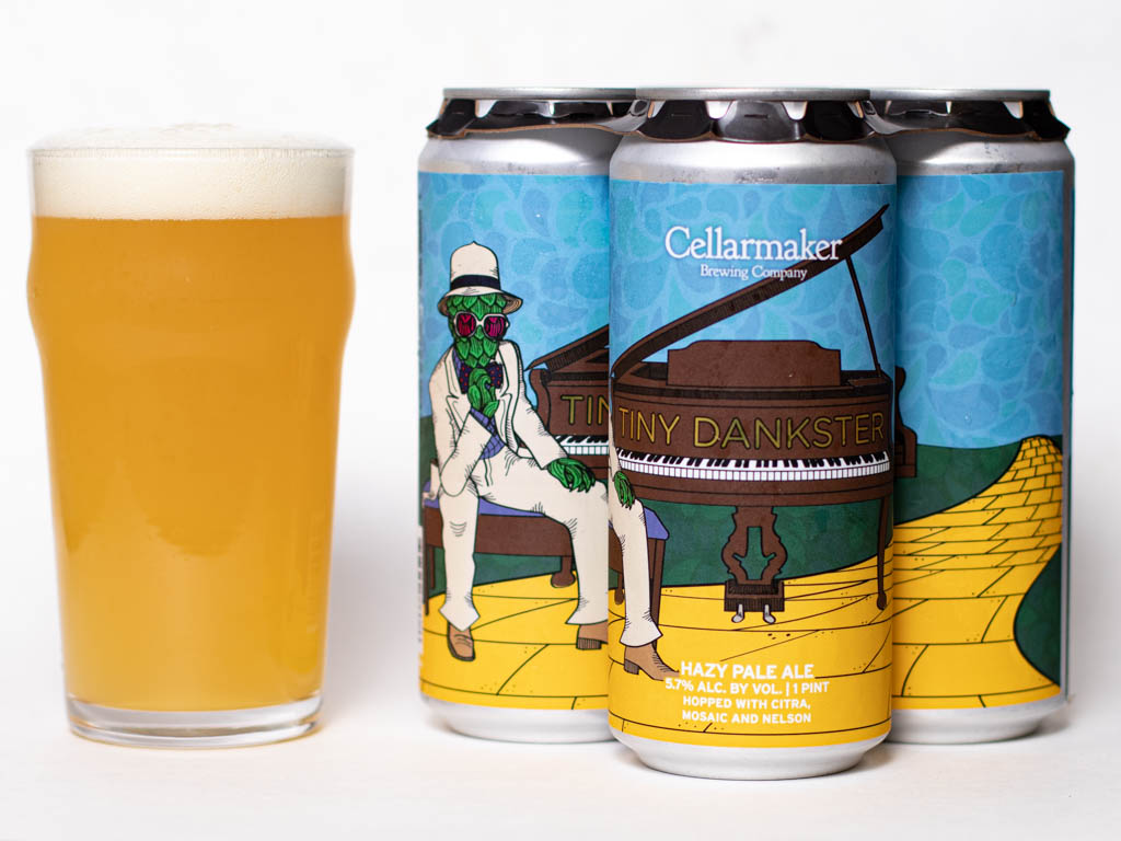 A Pint Glass Of Hazy Pale Ale Sits Next To Four Cans Of Cellarmaker Brewing Company'S Tiny Dankster. The Cans Feature Colorful Artwork Of A Frog In A Suit Playing Piano On A Yellow Brick Road.