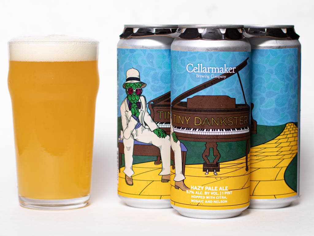 A Pint Glass Of Hazy Pale Ale Sits Next To Four Cans Of Cellarmaker Brewing Company'S Tiny Dankster. The Cans Feature Colorful Artwork Of A Frog In A Suit Playing Piano On A Yellow Brick Road.
