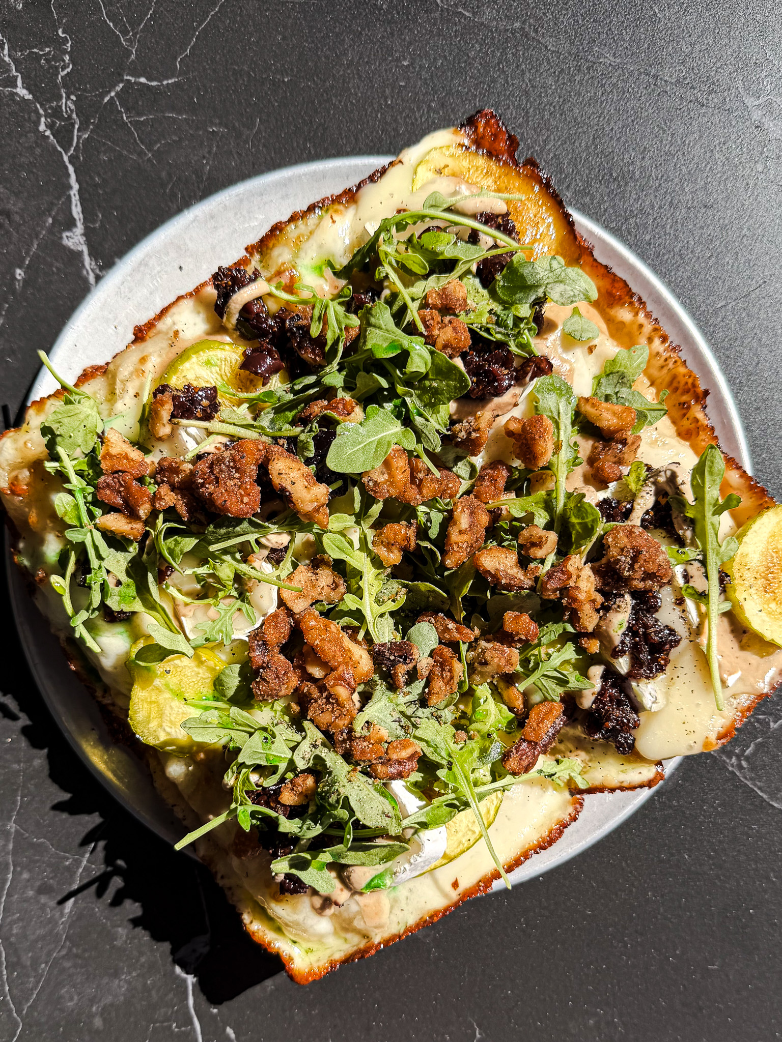 A Rectangular Pizza Topped With Arugula, Crumbled Sausage, Cheese, And Thinly Sliced Vegetables, Served On A Round Plate Atop A Dark, Marbled Surface.