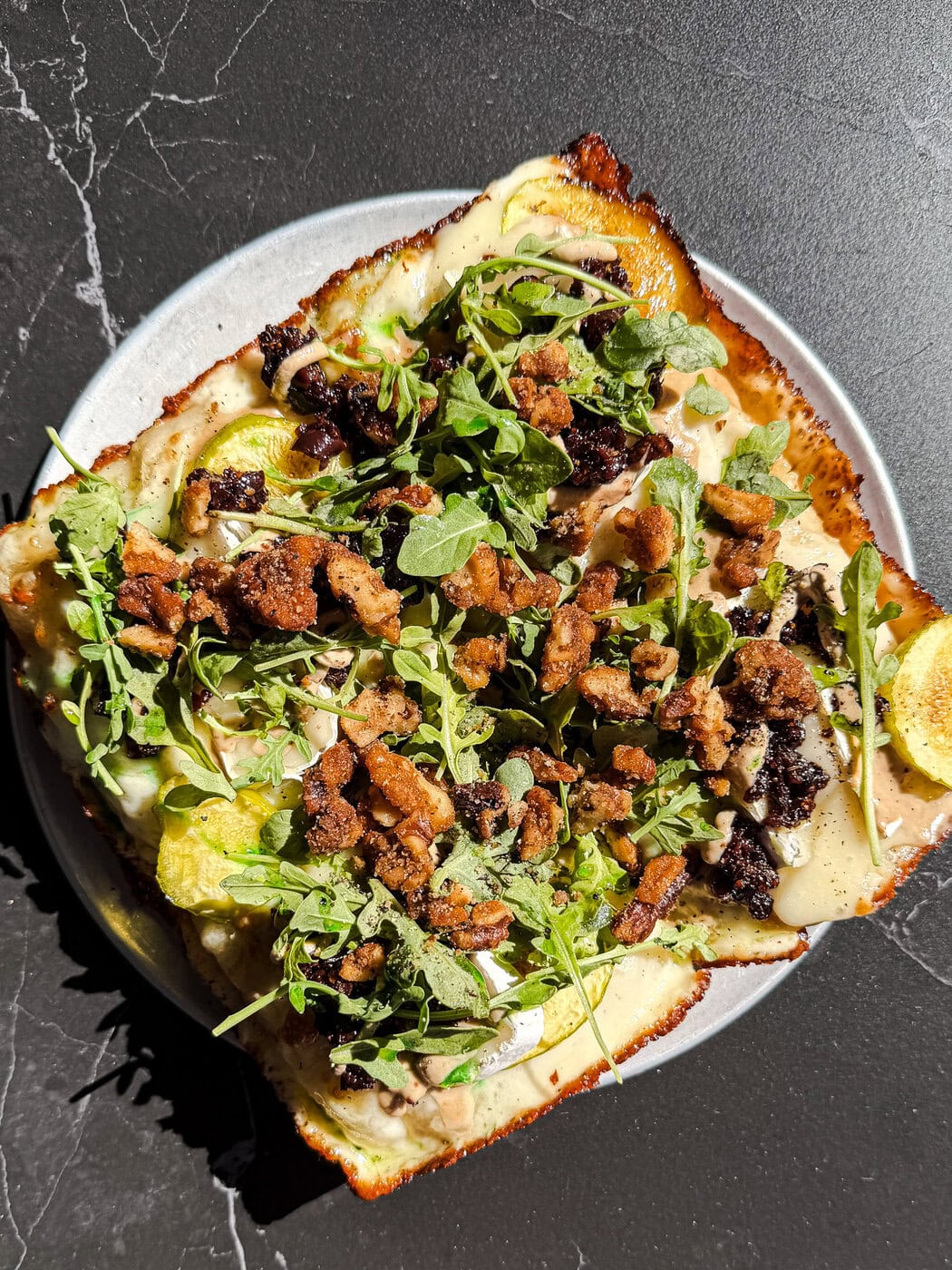 A Rectangular Pizza Topped With Arugula, Crumbled Sausage, Cheese, And Thinly Sliced Vegetables, Served On A Round Plate Atop A Dark, Marbled Surface.