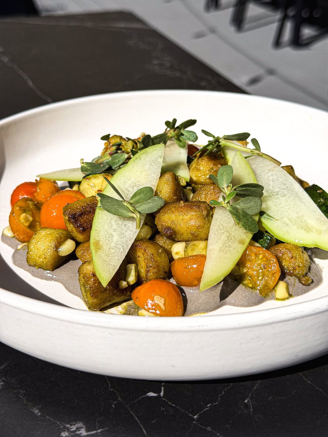 A White Bowl With Crispy Gnocchi, Cherry Tomatoes, Sliced Cucumber, Fresh Greens, Corn, And A Creamy Sauce, Served On A Dark Table.