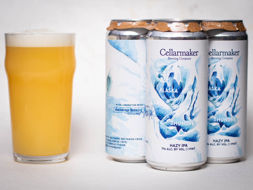 A Hazy, Light Yellow Beer In A Glass Sits Next To Three Tall Cans Labeled &Amp;Quot;Cellarmaker Alaska Thunder Hazy Ipa&Amp;Quot; With Blue And White Artwork Featuring Waves And Mountains.