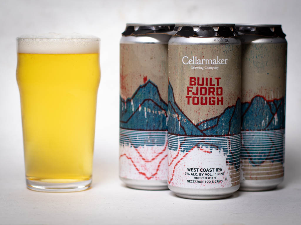 A Pint Glass Of Hazy Golden Beer Sits Next To Three Cans Labeled “Cellarmaker Brewing Company Built Fjord Tough West Coast Ipa” With Abstract Blue And Red Mountain Artwork On The Cans.