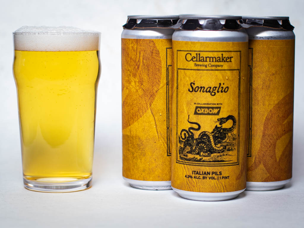 A Pint Glass Of Pale Beer Sits Next To Four Cans Of Cellarmaker Sonaglio Italian Pils, Featuring A Yellow Label With A Snake Illustration.