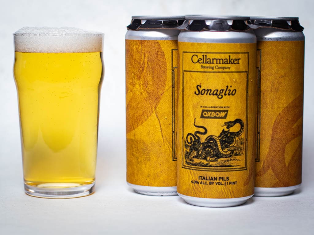 A Pint Glass Of Pale Beer Sits Next To Four Cans Of Cellarmaker Sonaglio Italian Pils, Featuring A Yellow Label With A Snake Illustration.