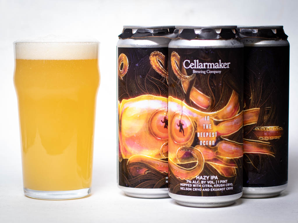 A Pint Glass Of Hazy Golden Beer Sits Next To Three Cans Labeled “Cellarmaker Brewing Company” With An Octopus And Diver Illustration And “In The Deepest Ocean Hazy Ipa” On The Front.