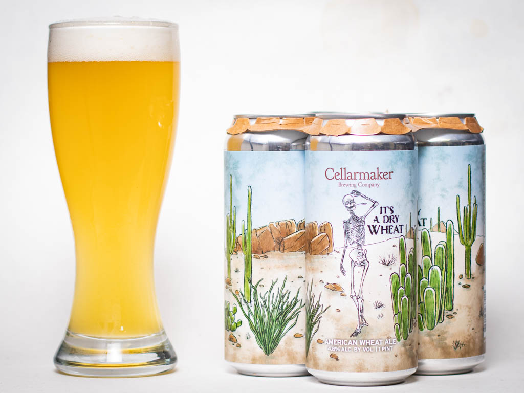A Tall Glass Of Hazy Pale Beer Stands Next To Three Cans Labeled &Amp;Quot;Cellarmaker It’s A Dry Wheat,&Amp;Quot; Featuring Desert Illustrations With Cacti And A Skeleton On The Can Art.