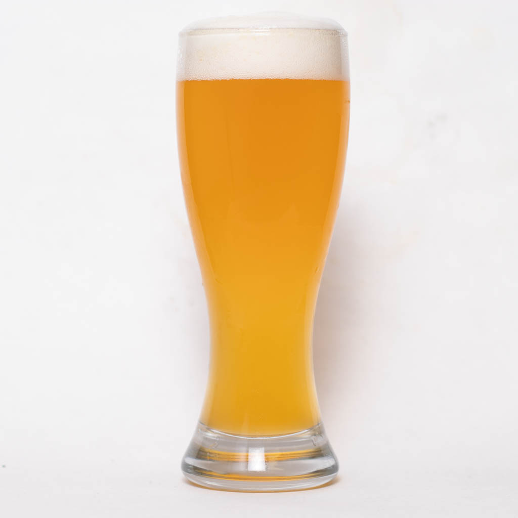 A Tall, Clear Glass Filled With Hazy, Golden Beer Topped With A Foamy White Head, Set Against A Plain White Background.