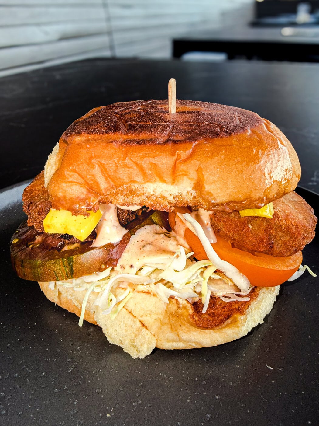 A Close-Up Of A Sandwich With Fried Chicken, Sliced Tomato, Shredded Lettuce, Pickles, Sauce, And A Toasted Bun, On A Black Plate.