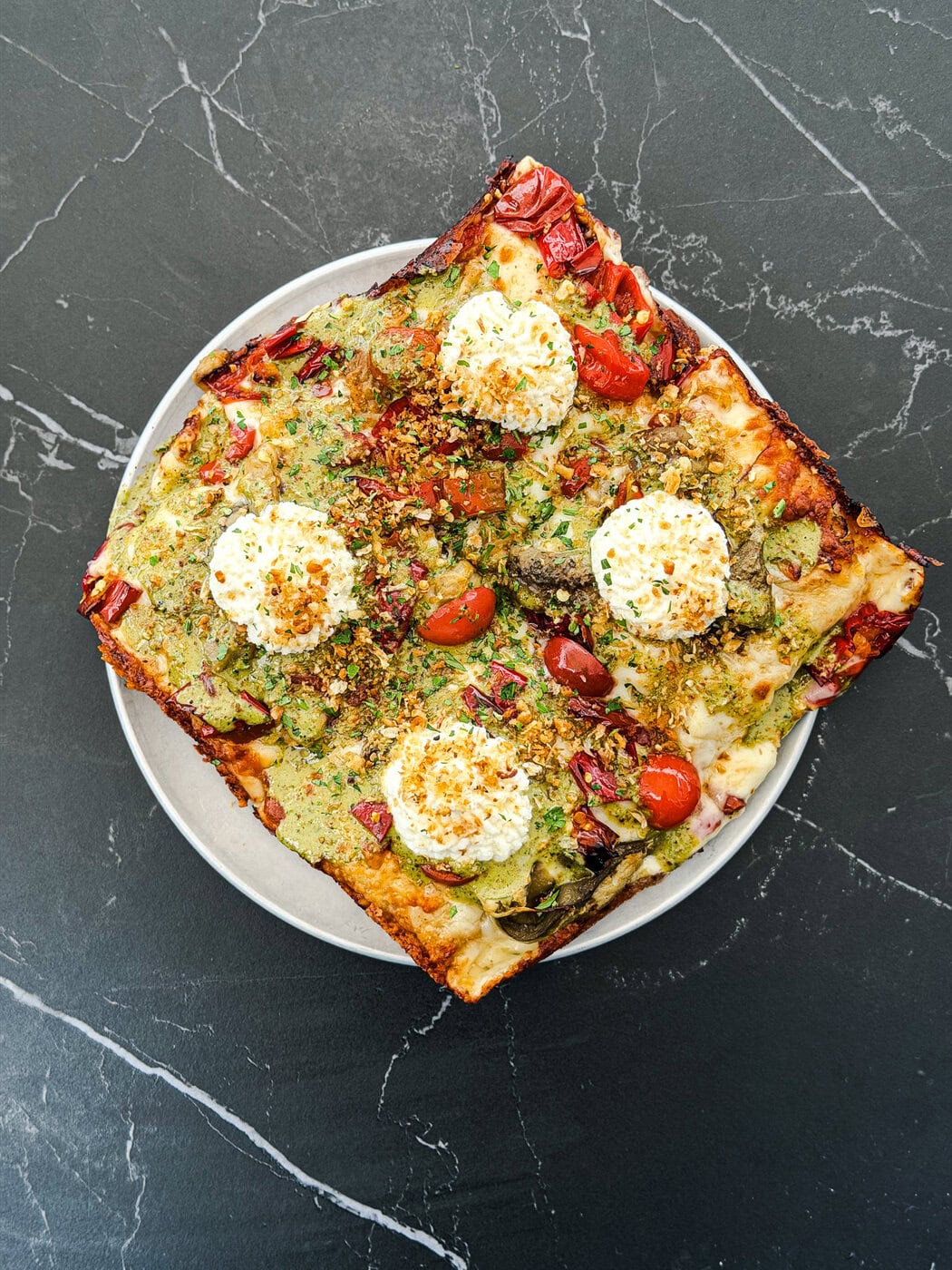 A Square Pizza On A Round Plate, Topped With Pesto, Ricotta Dollops, Cherry Tomatoes, Red Peppers, And Herbs, Sits On A Dark Marble Surface.