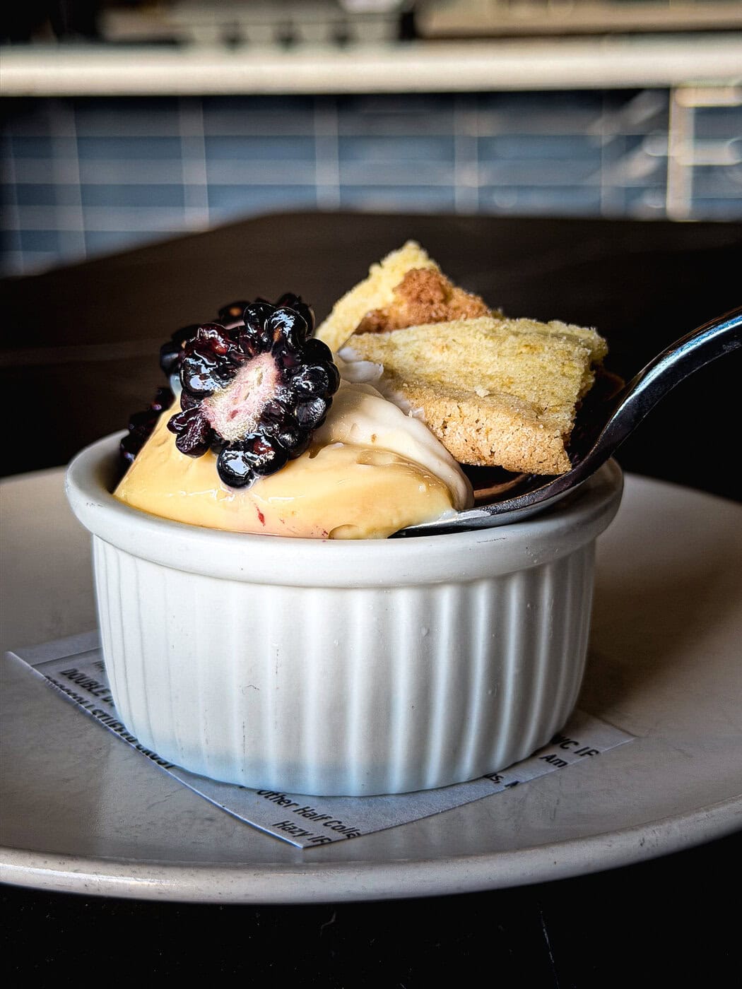 A Small White Ramekin Filled With Creamy Dessert, Topped With A Fresh Blackberry And Two Pieces Of Crumbly Biscuit, Sits On A White Plate With A Spoon.