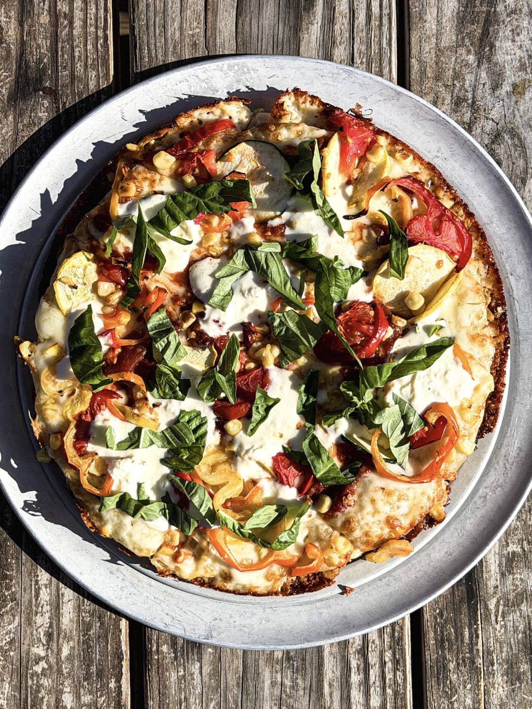 A Homemade Veggie Pizza Topped With Cheese, Tomato Slices, Bell Peppers, Zucchini, And Fresh Basil Sits On A Metal Tray Over A Rustic Wooden Table.