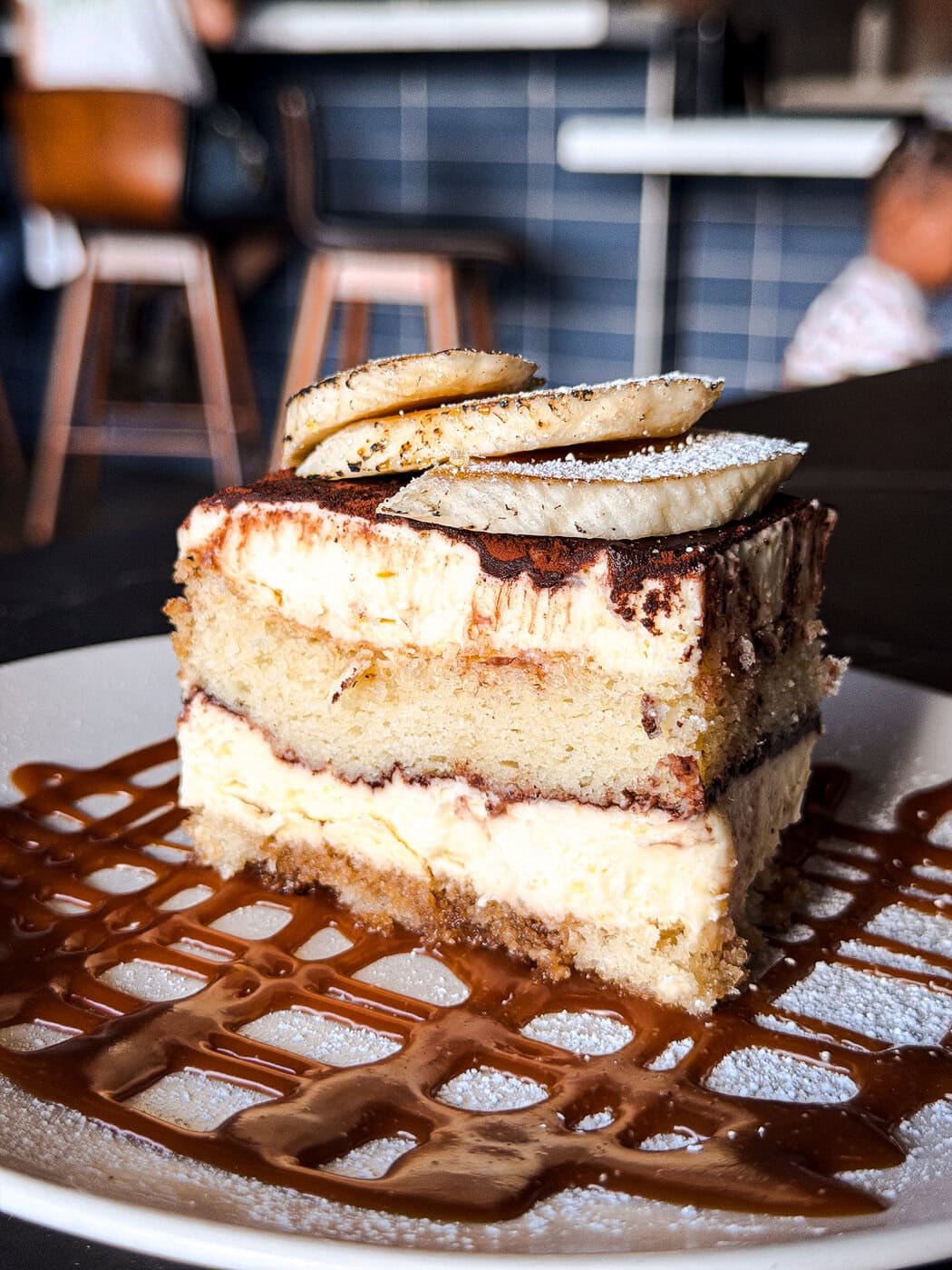 A Slice Of Layered Tiramisu Topped With Banana Slices Sits On A White Plate, Drizzled With Caramel Sauce. A Blurred Background Shows Chairs And Tiled Walls In A Cafe Setting.