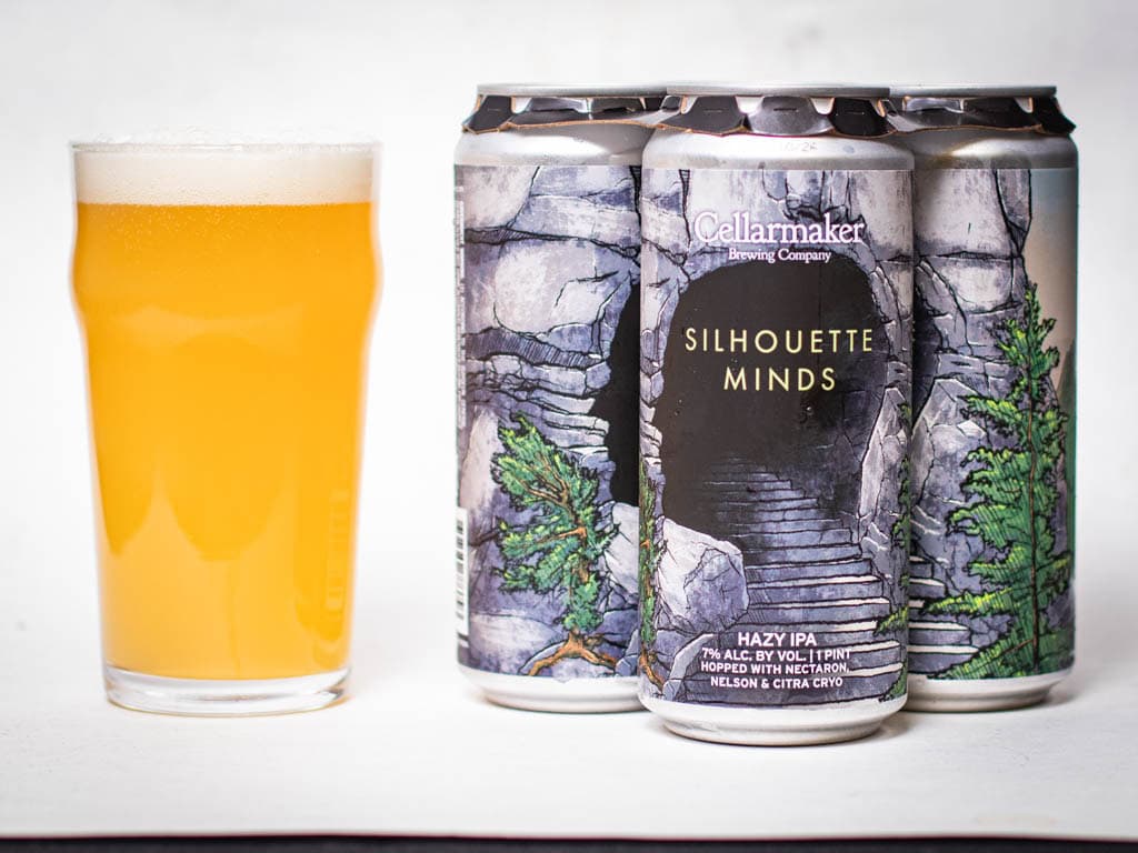 A Glass Of Hazy Yellow Beer Sits Next To Three Cans Of Cellarmaker Brewing’s Silhouette Minds Ipa, Featuring Artwork Of Stone Stairs And Pine Trees On The Labels.