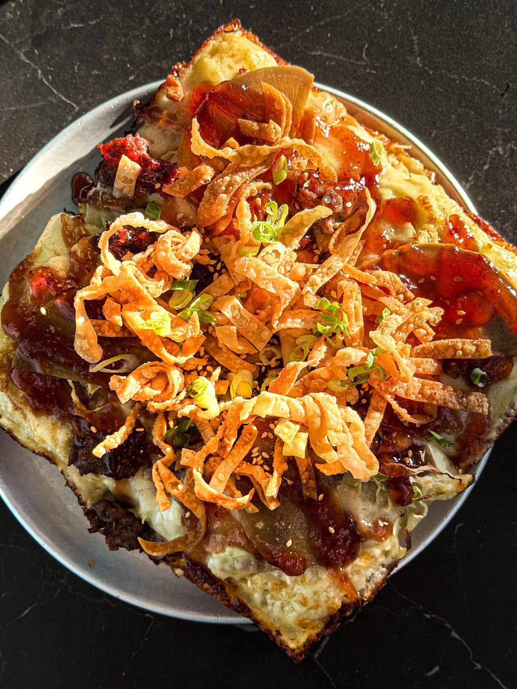 A Close-Up Of A Square Slice Of Pizza Topped With Crispy Fried Onions, Green Onions, Sauce, And Cheese On A Round Plate, Set On A Dark Surface.