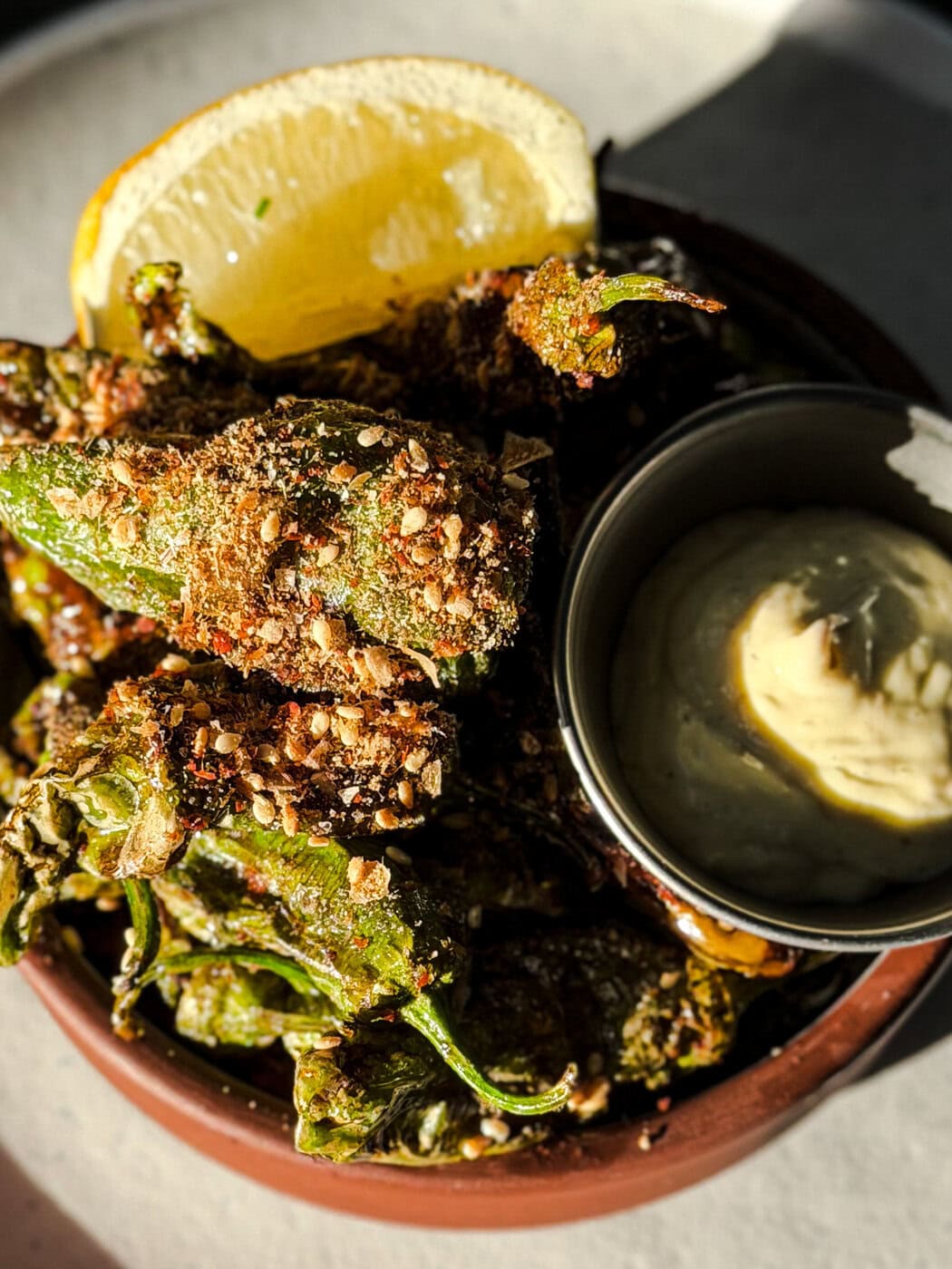 A Bowl Of Blistered Shishito Peppers Topped With Seasoning, Served With A Lemon Wedge And A Side Of Creamy Dipping Sauce.