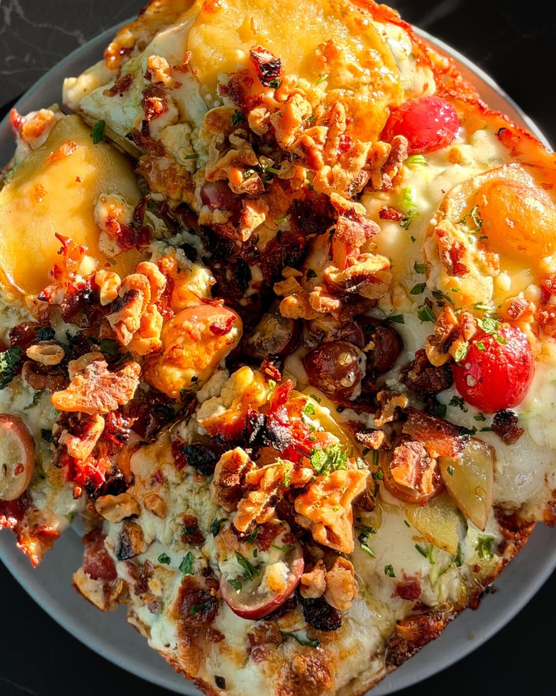 A Close-Up Of A Pizza Topped With Melted Cheese, Cherry Tomatoes, Walnuts, Herbs, And Sun-Dried Tomatoes On A White Plate In Sunlight.
