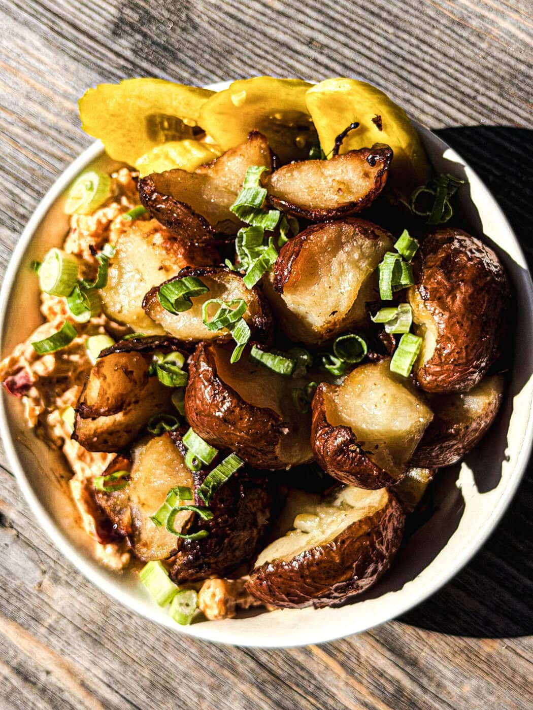 A bowl of roasted potatoes topped with chopped green onions, served with yellow pickles and a creamy, chunky dip, on a wooden surface.